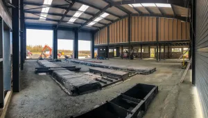 Interior of a hot rolled steel building, tall roof and open space with open doors leading out into a parking area.