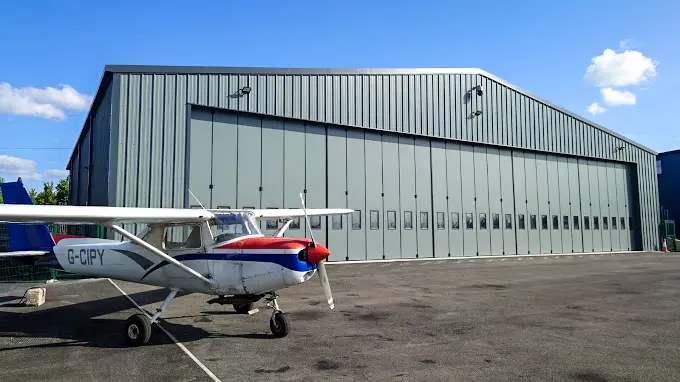 Large steel aircraft hangar in grey with concertina doors and a small aeroplane in the foreground at Elstree Aerodrome