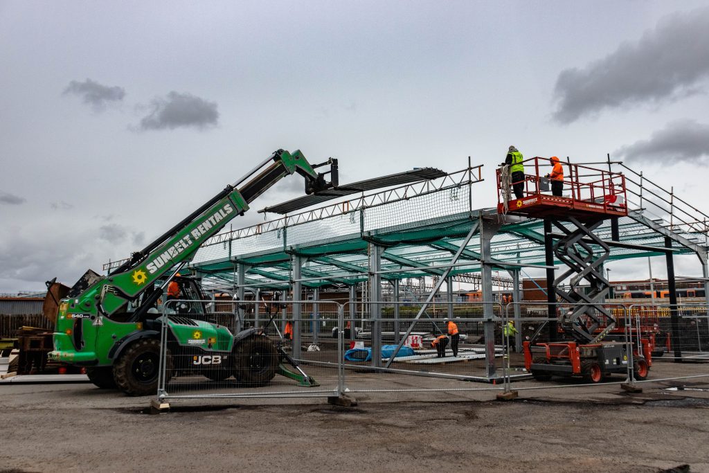 steel train shed building under construction with steel workers on a scissor lift and a telehandler lifting cladding